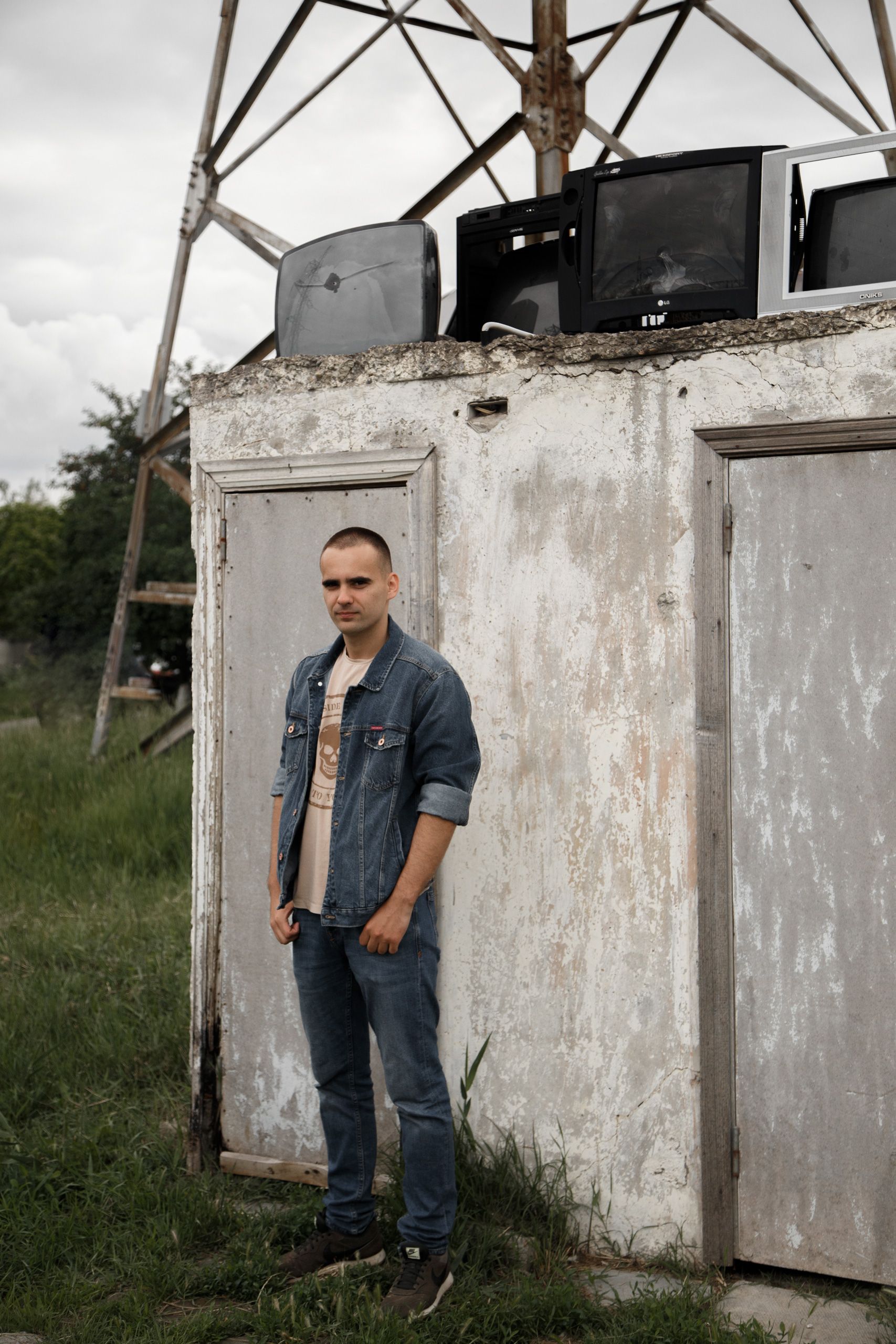 Young man in a denim outfit poses next to weathered building with old TVs outdoors.