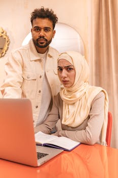 Two colleagues, one in a hijab, intently reviewing work on a laptop in an office setting.