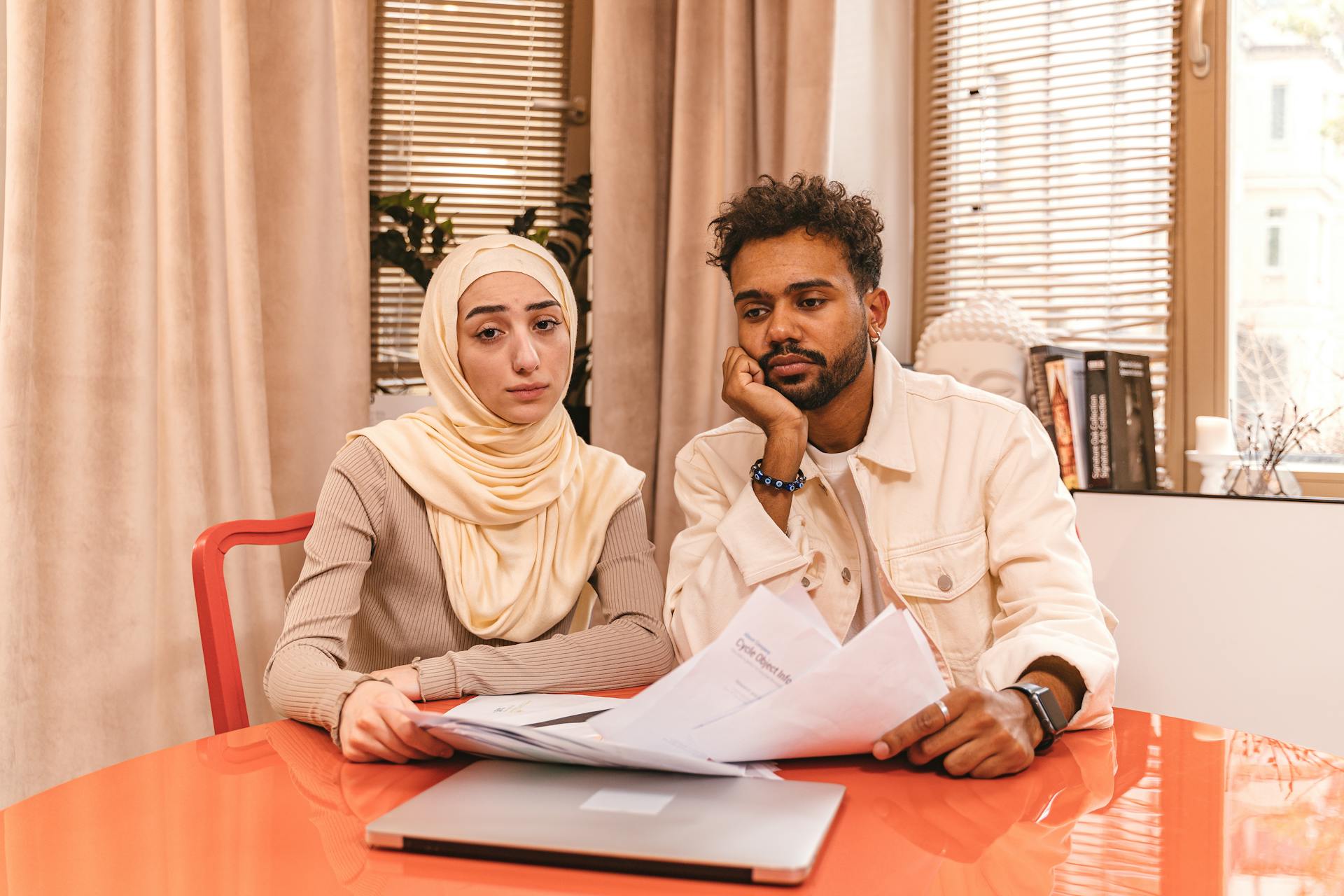 A couple in casual attire sits indoors at a table reviewing financial documents with a laptop.