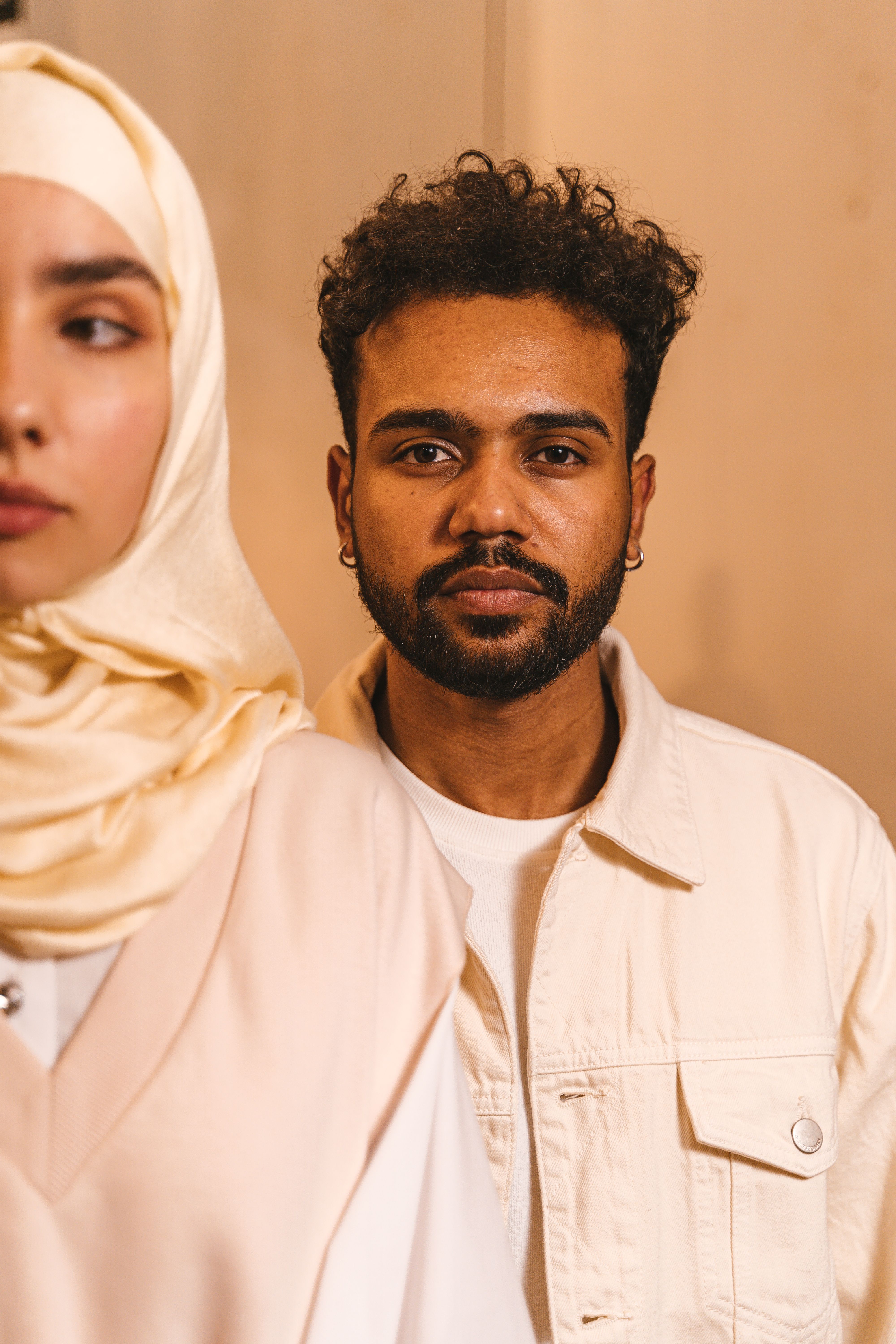 A man with a beard in a light beige jacket stands in an indoor setting next to a woman.