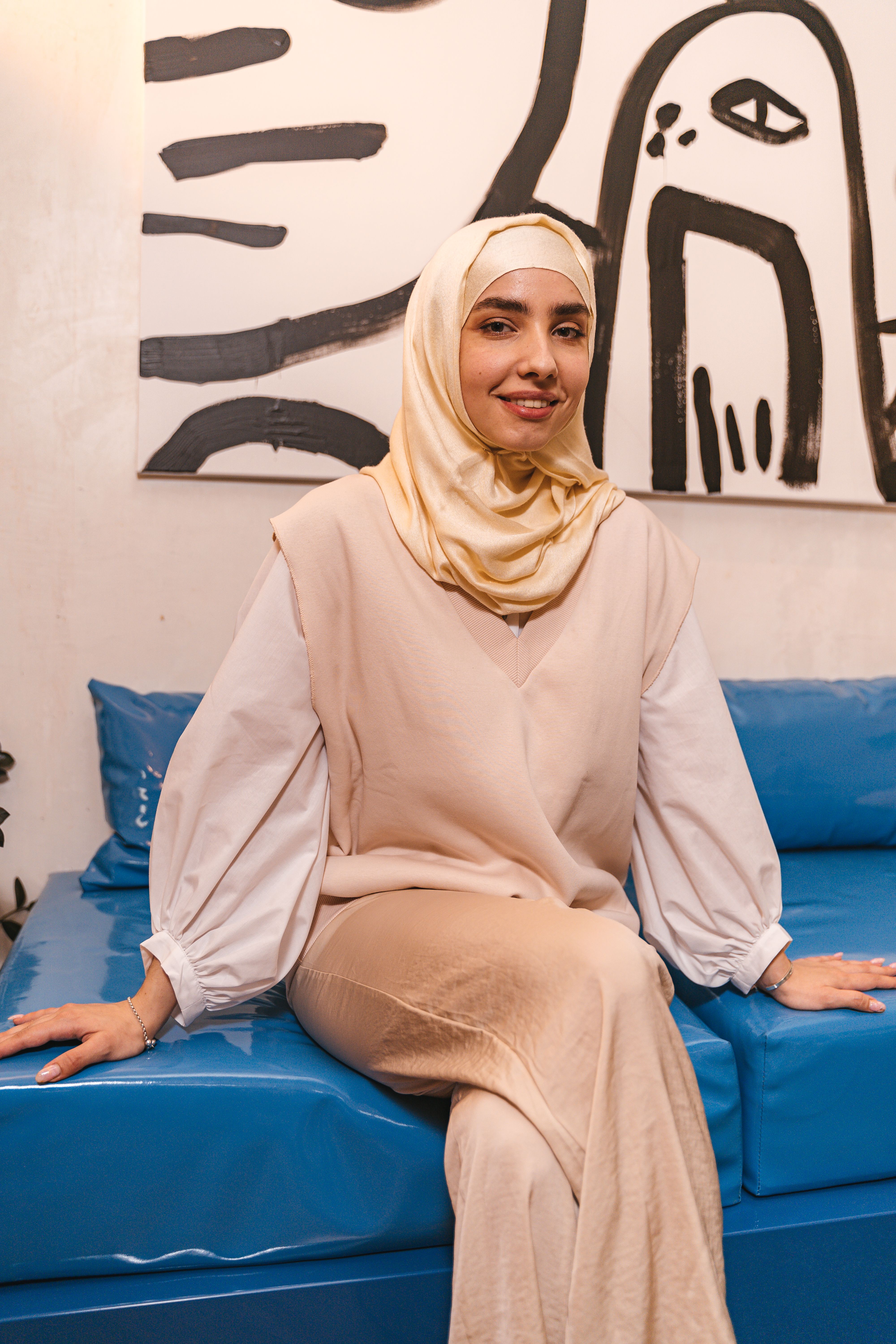 A woman in a hijab sitting on a blue sofa, smiling indoors.