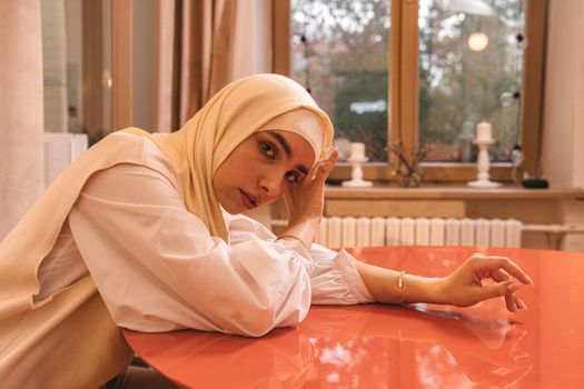 Portrait of a woman in a hijab sitting at a table indoors, creating a tranquil scene.