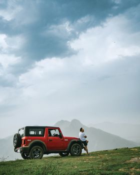 A man leans on a red SUV with a mountainous backdrop under cloudy skies.