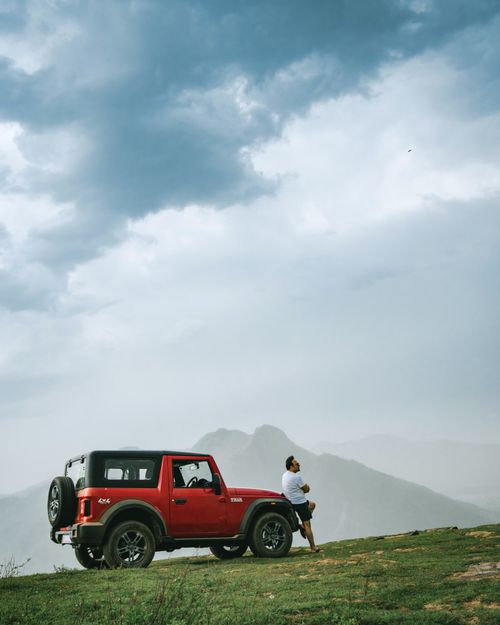 Free A man leans on a red SUV with a mountainous backdrop under cloudy skies. Stock Photo