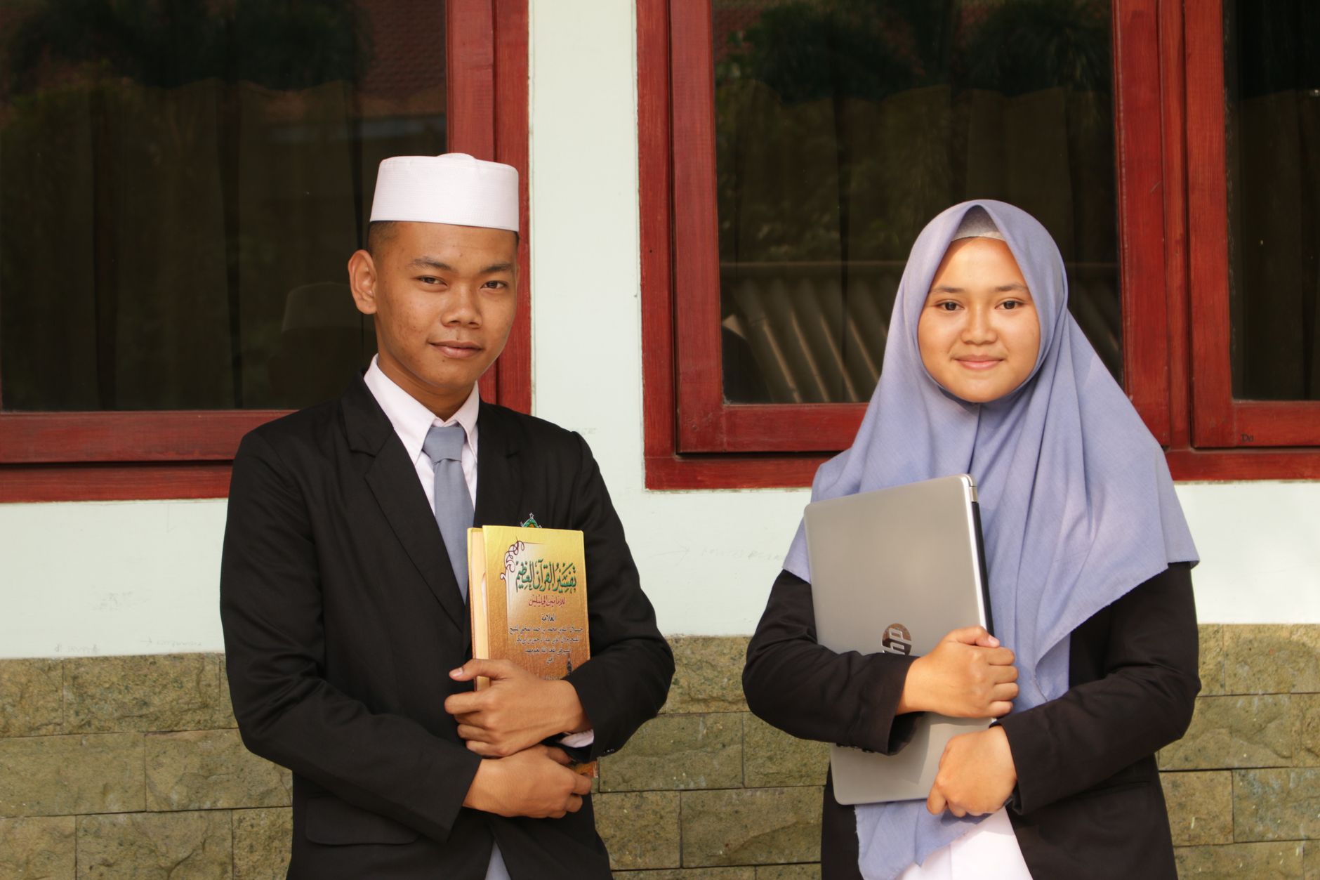 Two Indonesian students in traditional attire standing outdoors with books and laptop.