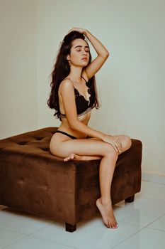 An adult woman in lingerie seated on a brown ottoman indoors with closed eyes.