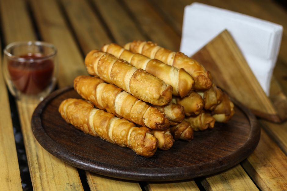 A pile of golden fried tequeños stacked on a plate, one broken open to show melted white cheese stretching from the center, served with a small bowl of guasacaca sauce.