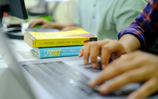 Hands typing on a laptop with design and problem-solving books in the background.