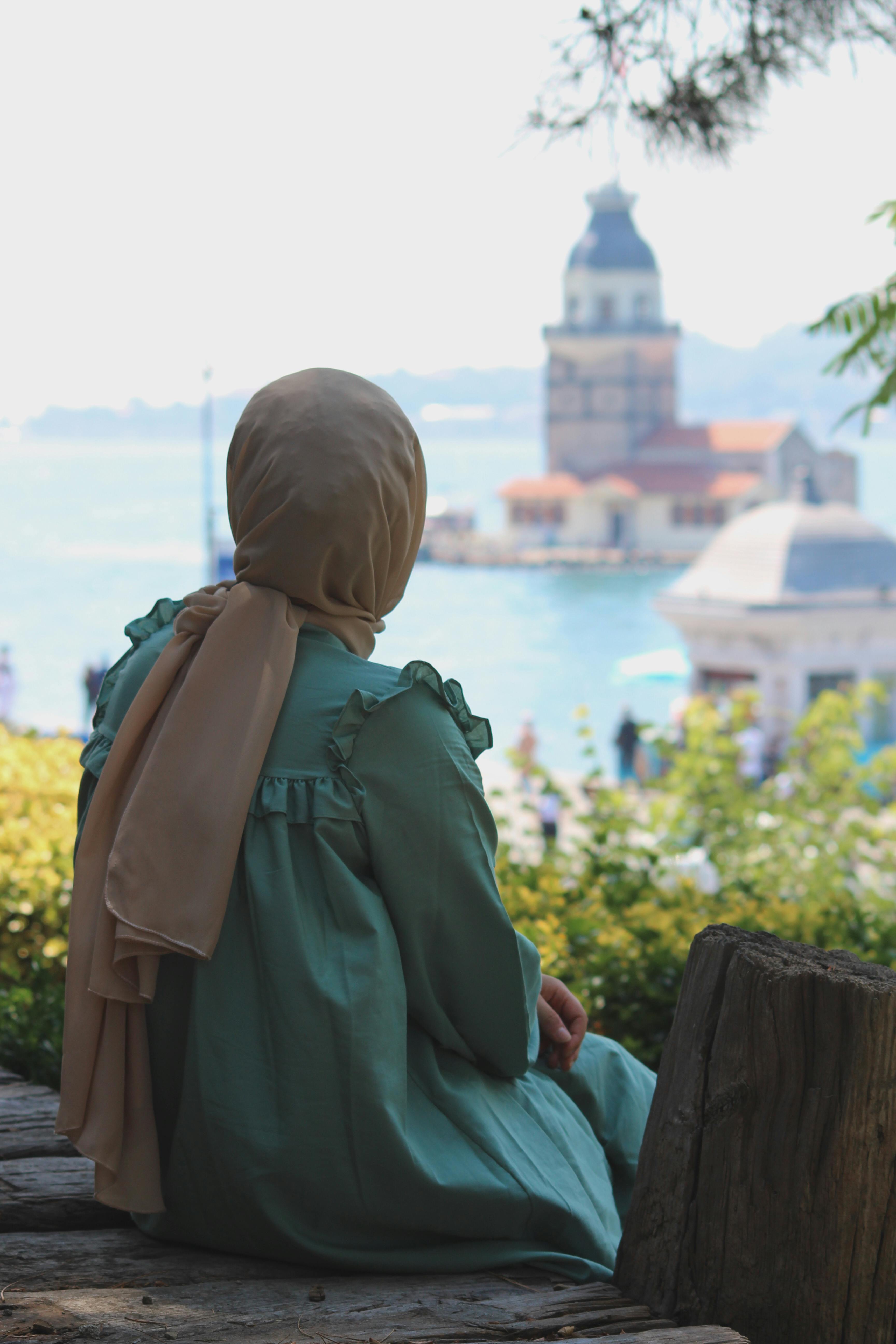 A woman in a hijab sits outdoors, gazing at a picturesque water scene with a distant historical tower.