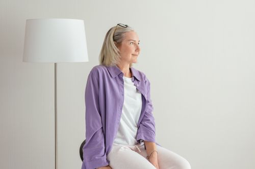 Free Portrait of a senior woman in casual attire, sitting indoors with a lamp beside her. Stock Photo