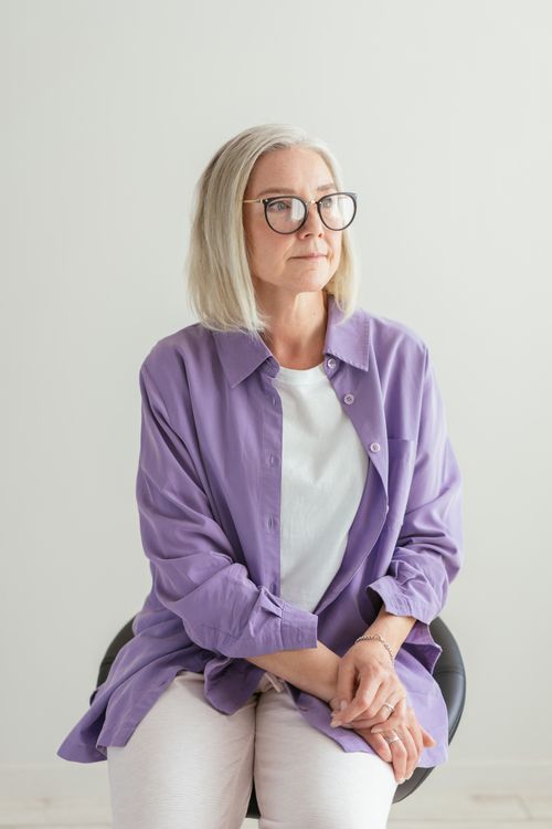 Free Elegant senior woman with glasses sitting indoors wearing a purple shirt, exuding a calm and thoughtful expression. Stock Photo