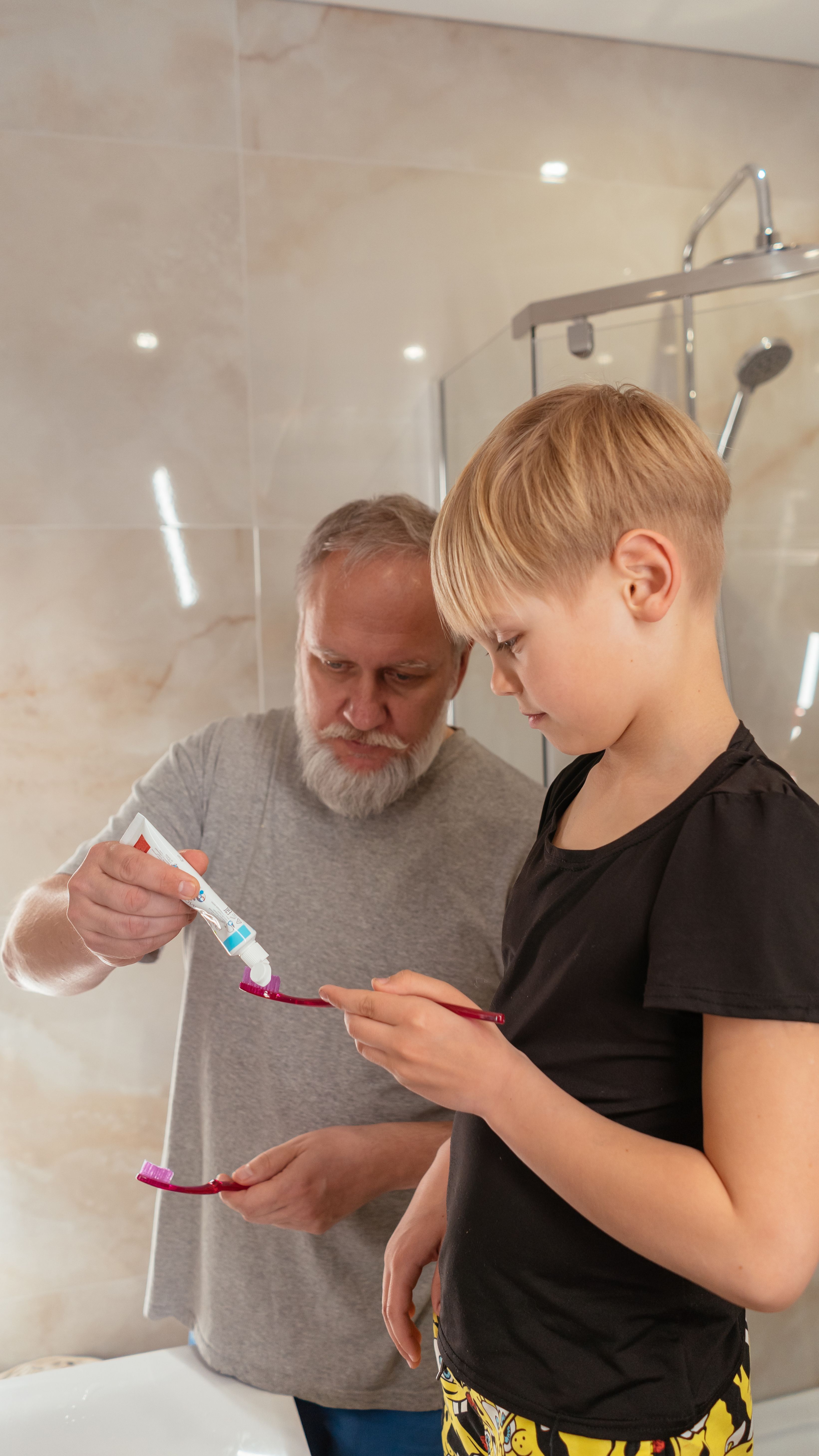 An elderly man helps a young boy with dental hygiene in a bathroom setting.