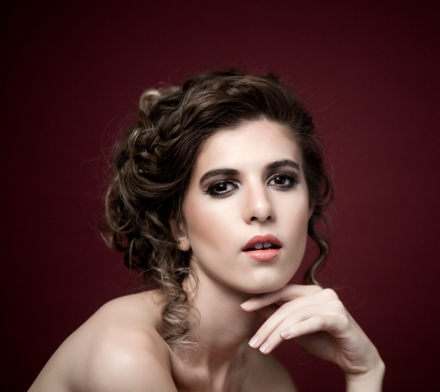 Close-up portrait of a woman with intricate hairstyle and makeup against a deep maroon background, exuding elegance and beauty.