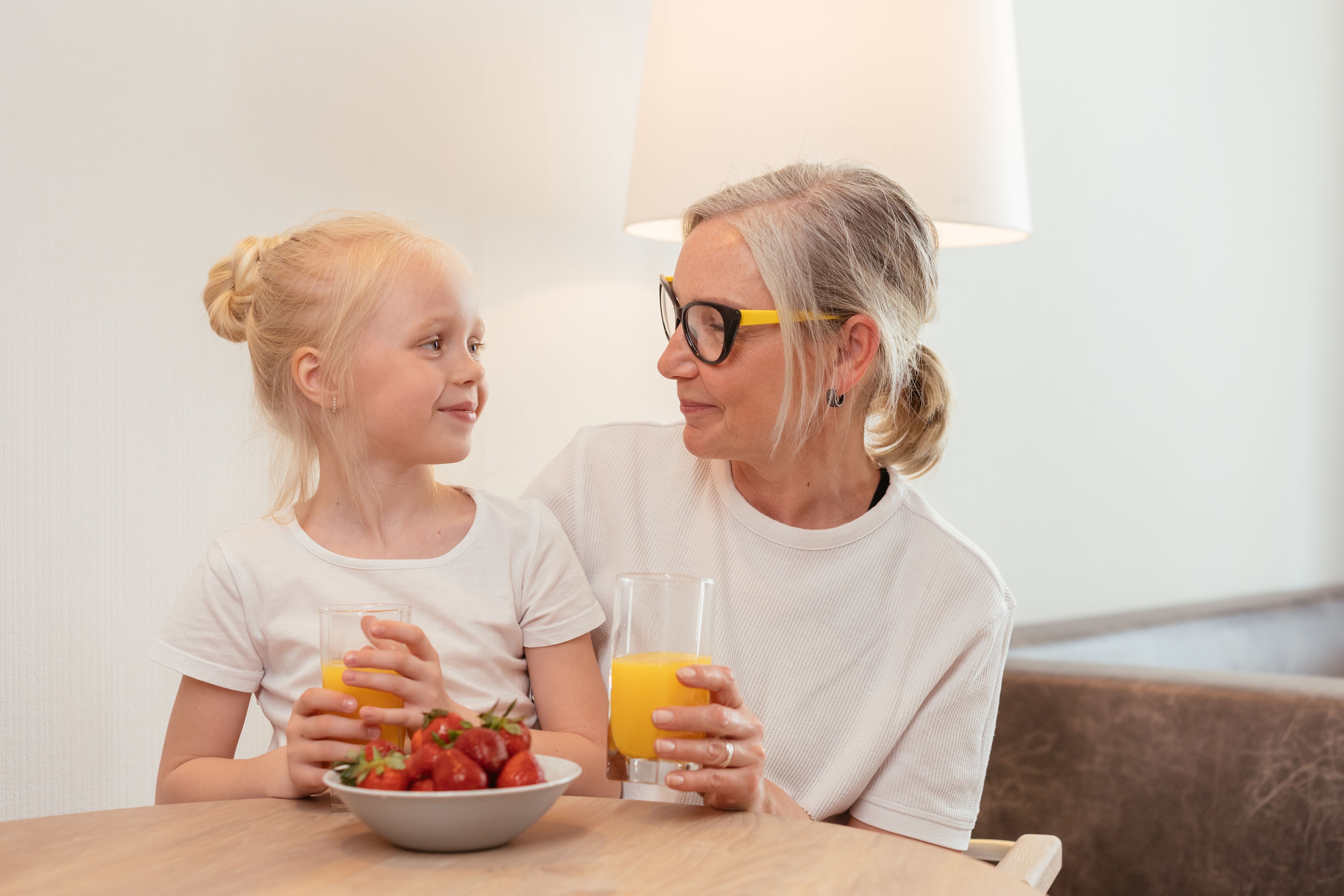 Elderly woman and young girl enjoying orange juice and strawberries at home, sharing a joyful moment together.