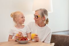 Elderly woman and young girl enjoying orange juice and strawberries at home, sharing a joyful moment together.