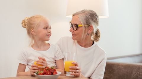 Elderly woman and young girl enjoying orange juice and strawberries at home, sharing a joyful moment together.