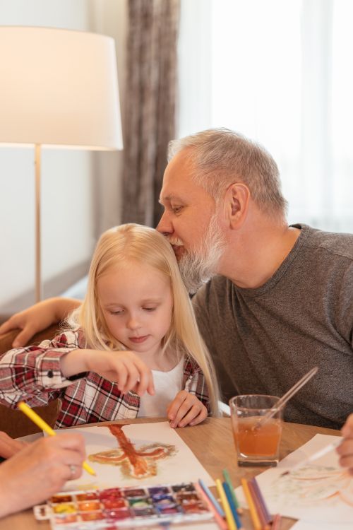 Free Grandfather helping granddaughter paint, showcasing family bonding and creativity at home. Stock Photo