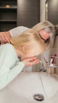 Elderly woman guiding young girl in washing hands at the sink for hygiene.