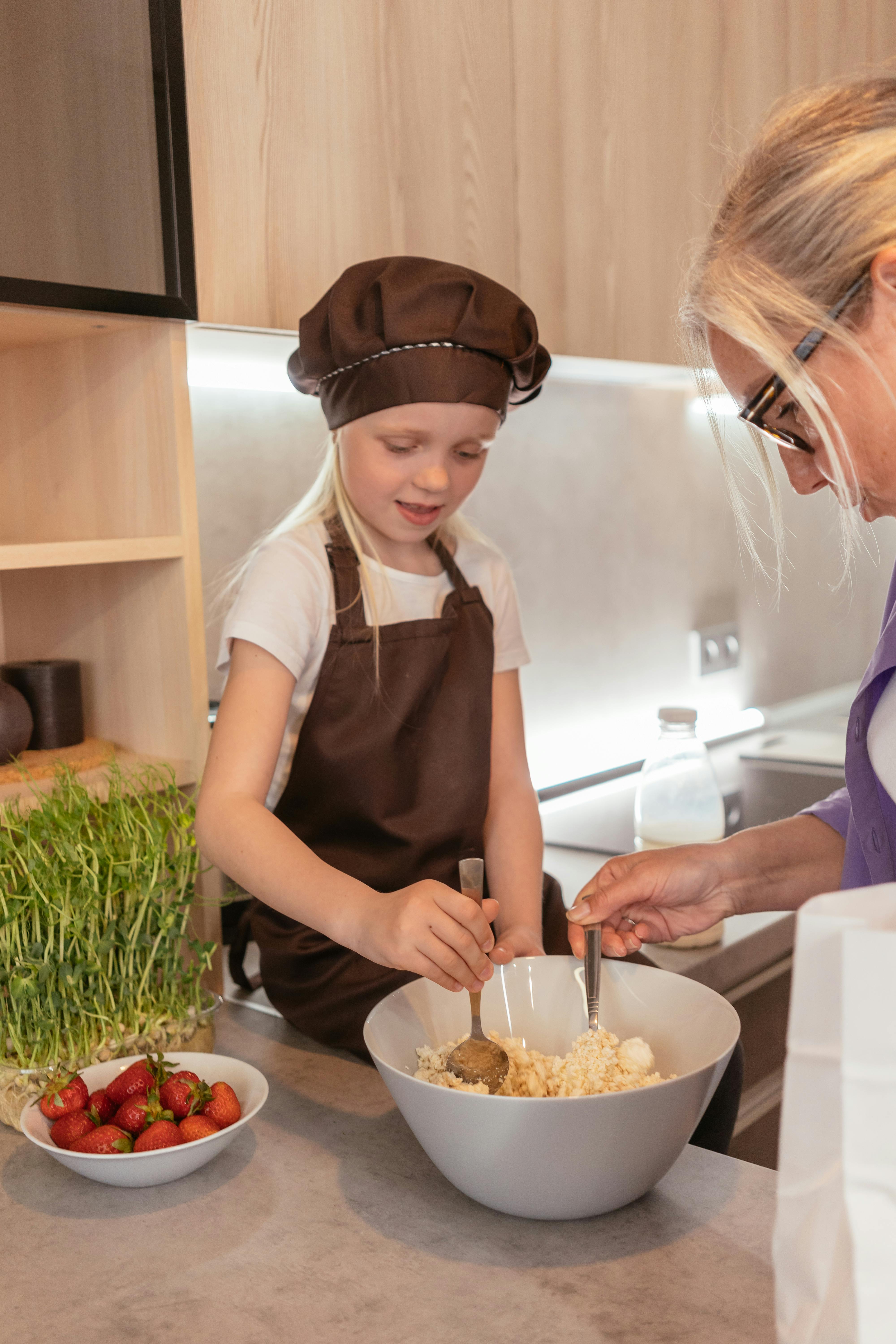 girl in brown apron with chef hat mixing food in a bowl beside a woman