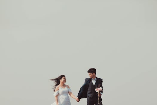 Young couple in wedding attire holding hands with a clear sky backdrop, symbolizing love and unity.