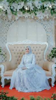 A beautiful Muslim bride sitting on an ornate sofa in a stunning gown and hijab, ready for her wedding day.