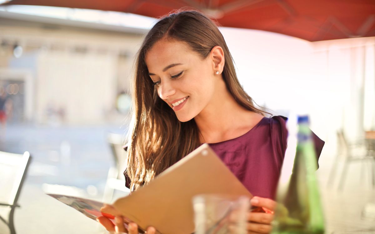A woman smiles as she reads a menu at an outdoor restaurant, enjoying a sunny day.