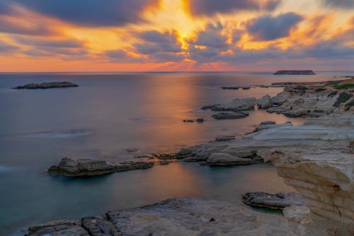 Shore with rough stones washed by calm sea against colorful sky with clouds at sundown time in nature on summer evening