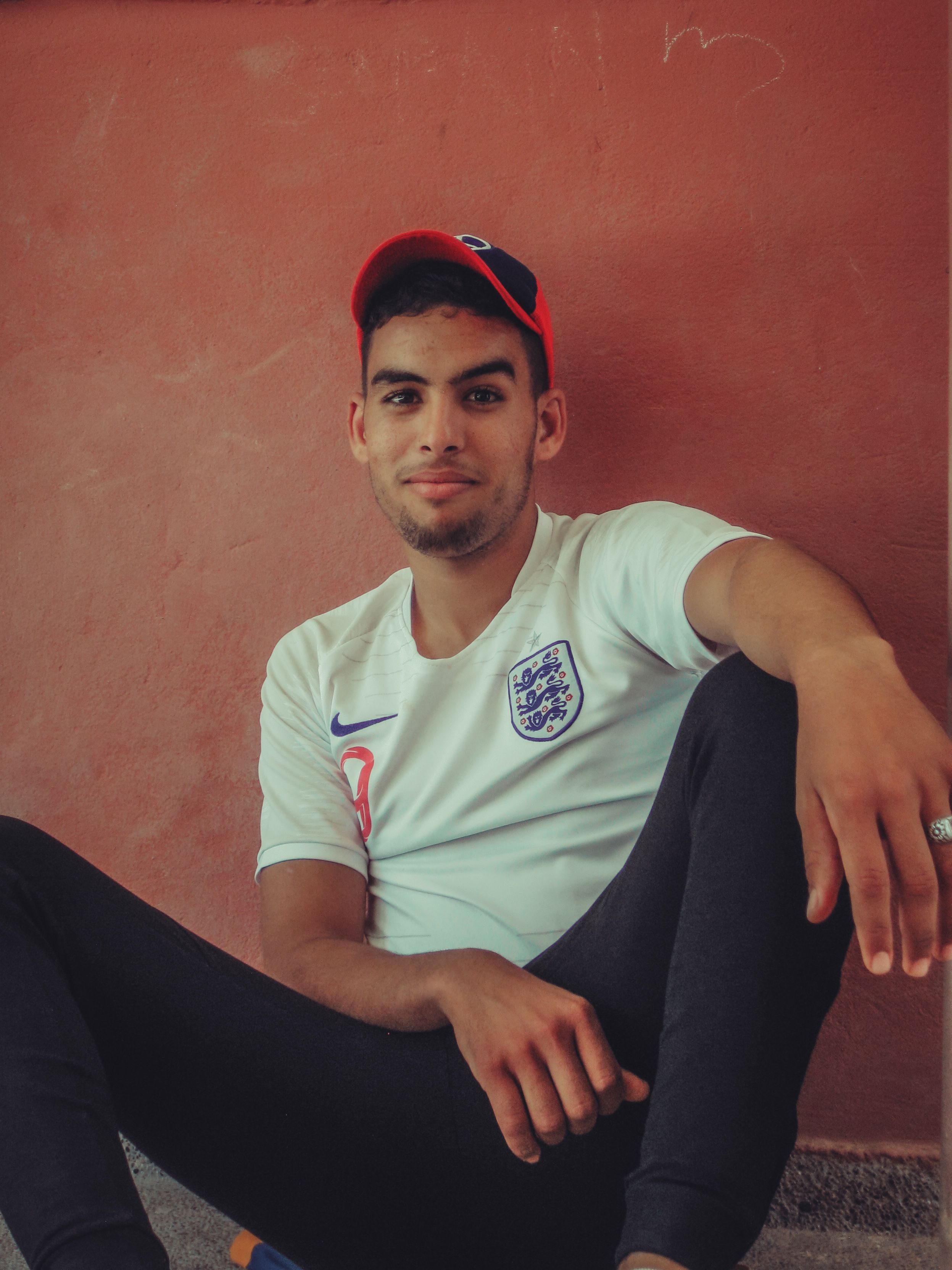 Young man in England jersey sitting against a brown wall, smiling casually.