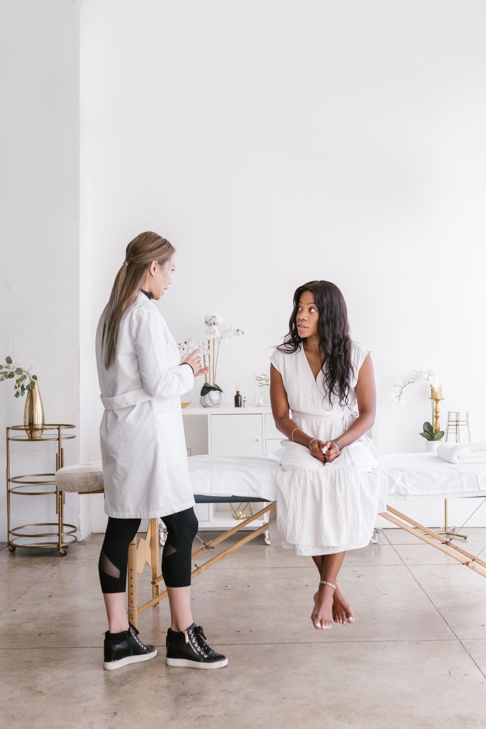 A therapist in a white coat stands beside a seated client in a bright, minimal treatment space, talking through her needs.