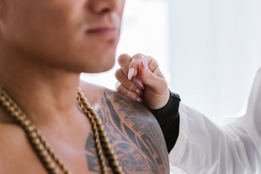 Close-up of tattooed man's shoulder, receiving acupuncture treatment by a practitioner.