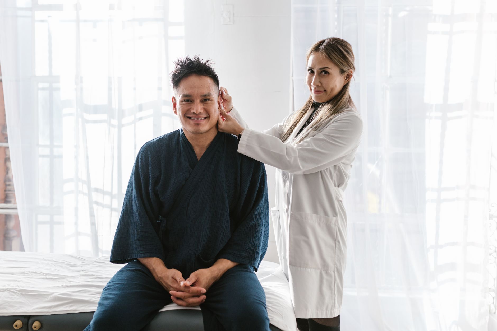 A doctor in a white coat examines a smiling patient indoors in a bright setting.