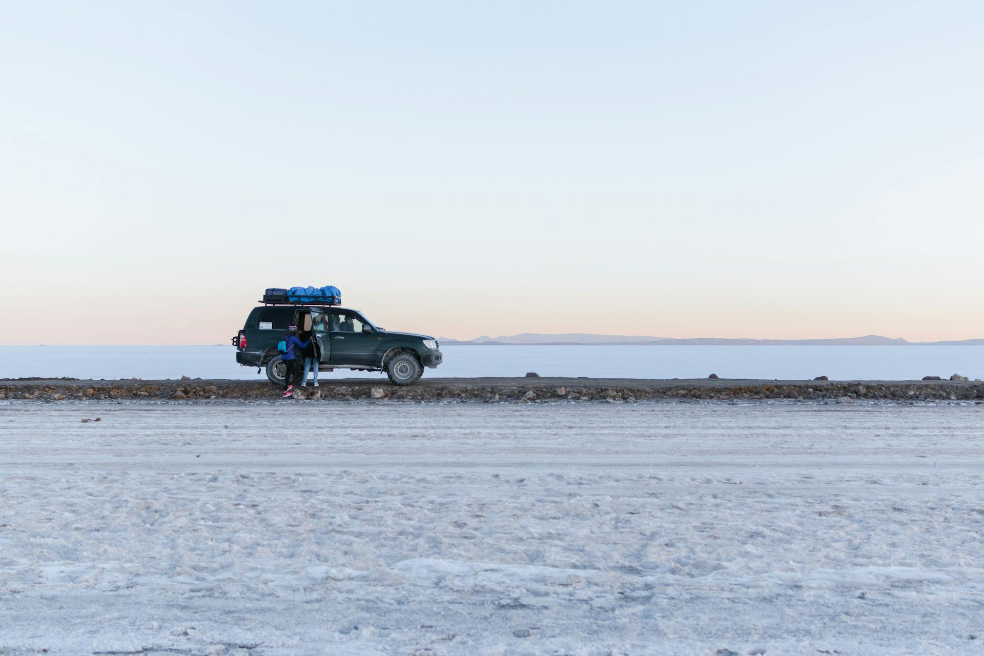 A 4x4 SUV with travelers in the vast, icy landscape of the Salar de Uyuni at sunrise.