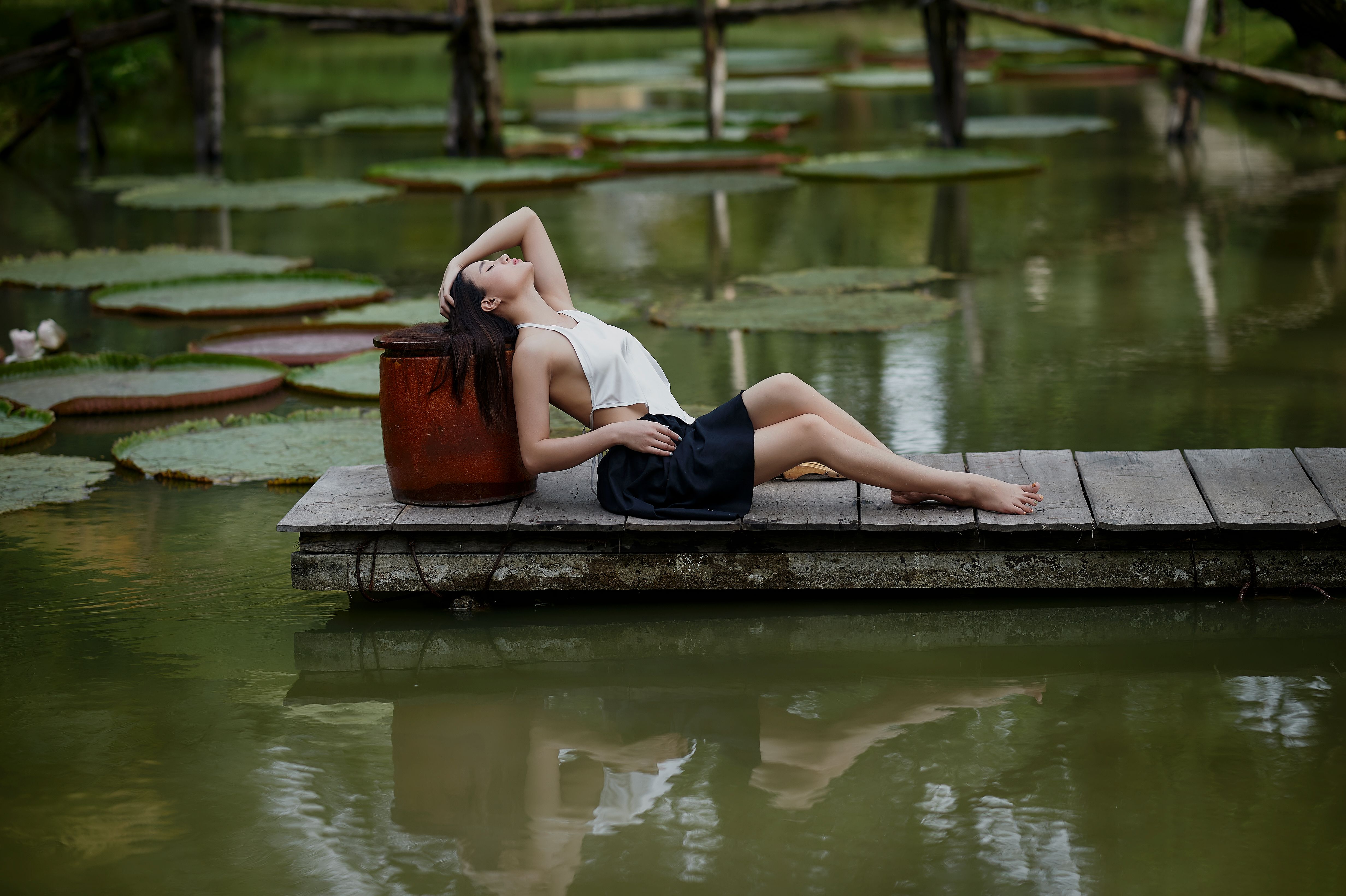 A serene woman relaxes on a dock beside a tranquil pond with lily pads, exuding peace and calm.