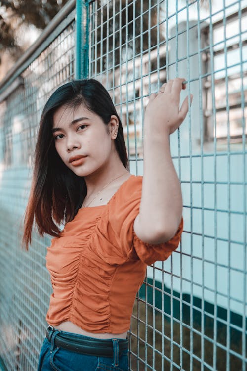 Free Woman in Orange Top Posing on Metal Fence Stock Photo