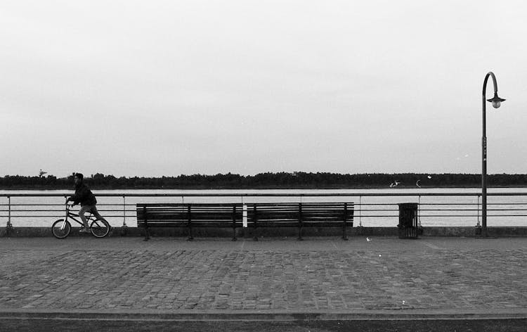 Black And White Photo Of A Kid Riding A Bike By The River