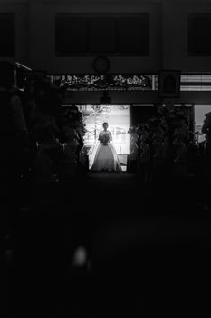 Black and white of unrecognizable bribe in elegant white wedding dress with flowers in hand standing in doorway in dark room