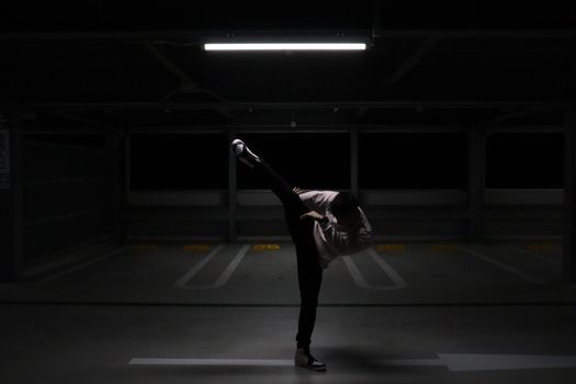 A man performs a high kick in an empty parking garage with dynamic lighting.