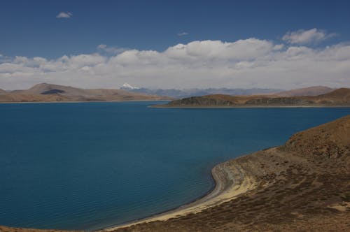 08 Days Kailash Mansarovar Tour via Lhasa | Fly-In Yatra Package 1 Free Breathtaking view of a lake surrounded by mountains under a blue sky in Tibet, China. Stock Photo