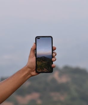 A hand holding a smartphone displaying a mountain view against a blurred landscape background.