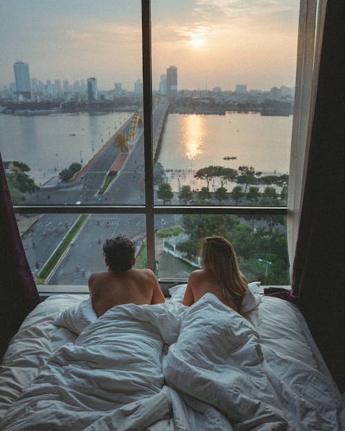 A Couple Lying on the Bed in Front of the Glass Window with City View