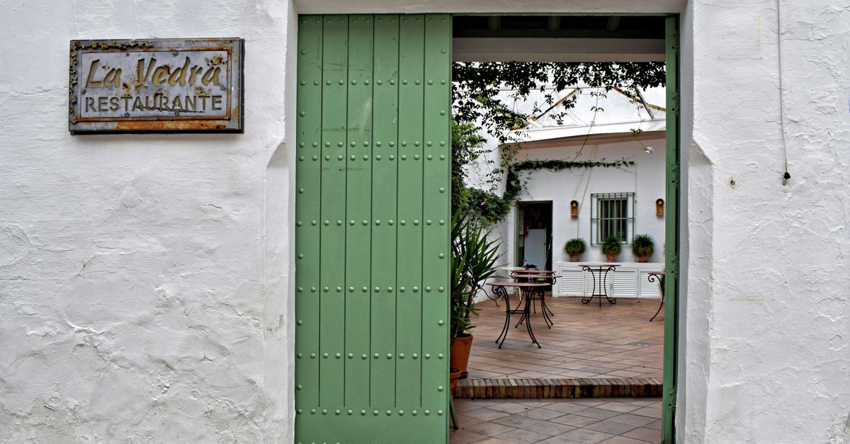 A picturesque doorway leading to a quaint Spanish café courtyard featuring rustic tables and plants.