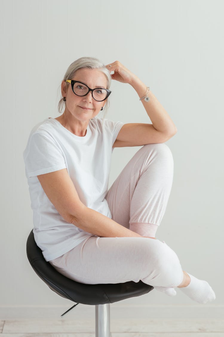 An Elderly Woman In White Shirt Smiling While Sitting On The Chair