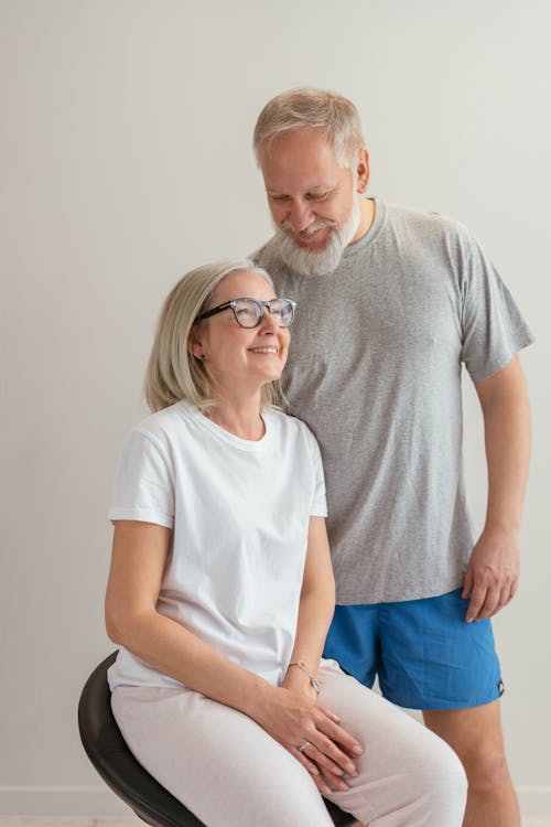 An Elderly Man Standing Beside the Woman in White Shirt Sitting on the Chair