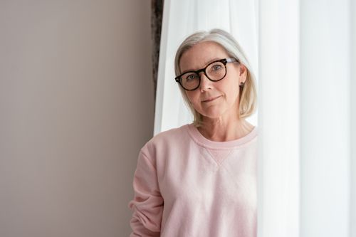 Free Elegant senior woman with glasses standing by a bright curtain indoors. Stock Photo
