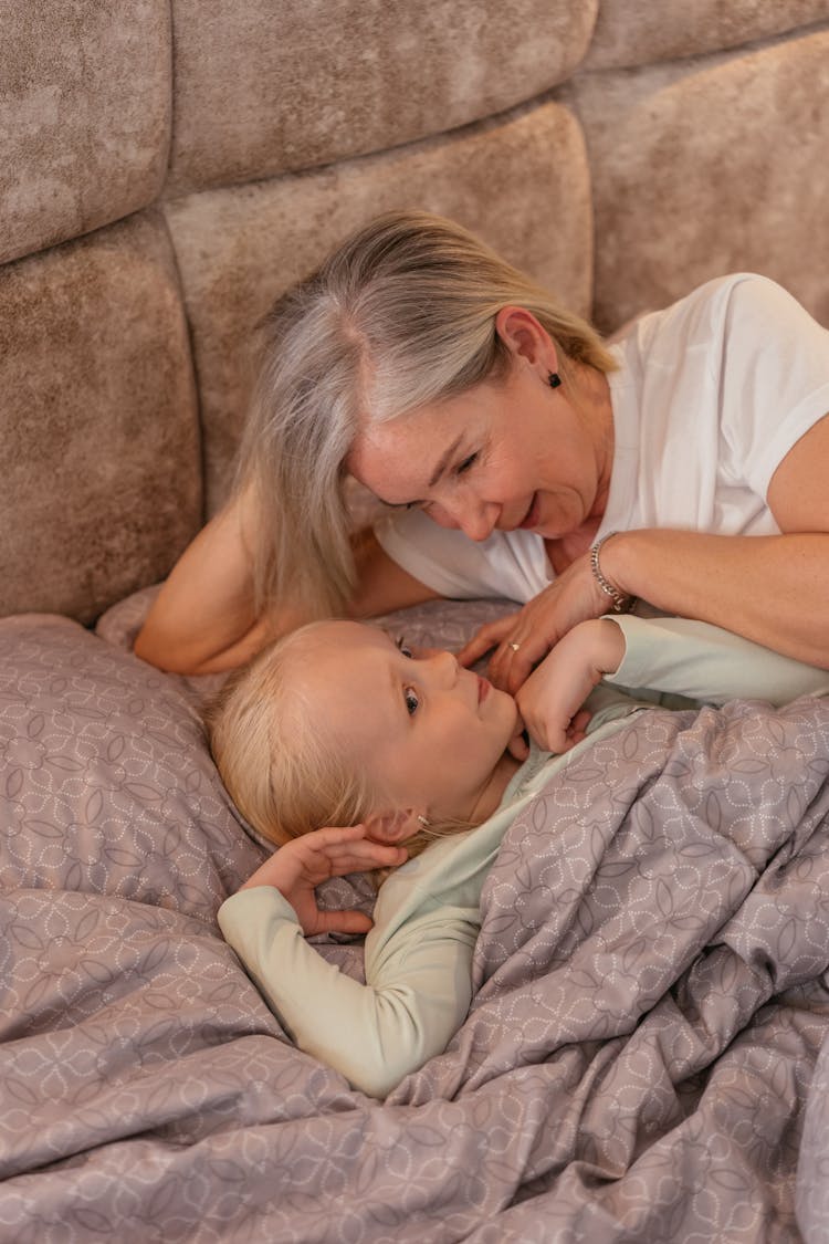 An Elderly Woman In White Shirt Talking To Her Granddaughter While Lying On The Bed