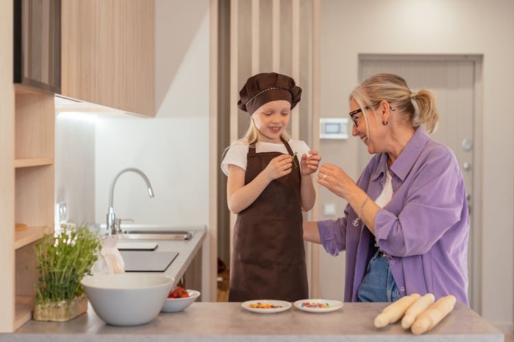 An Elderly Woman In Purple Long Sleeves Talking To Her Grandchild Wearing Apron And Brown Cap