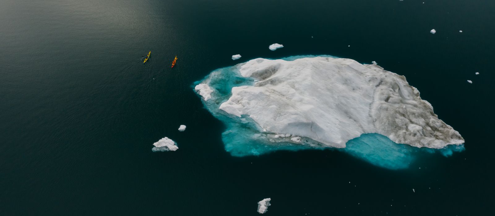 Kayaks paddling past a large blue-white iceberg in cold glacial water.