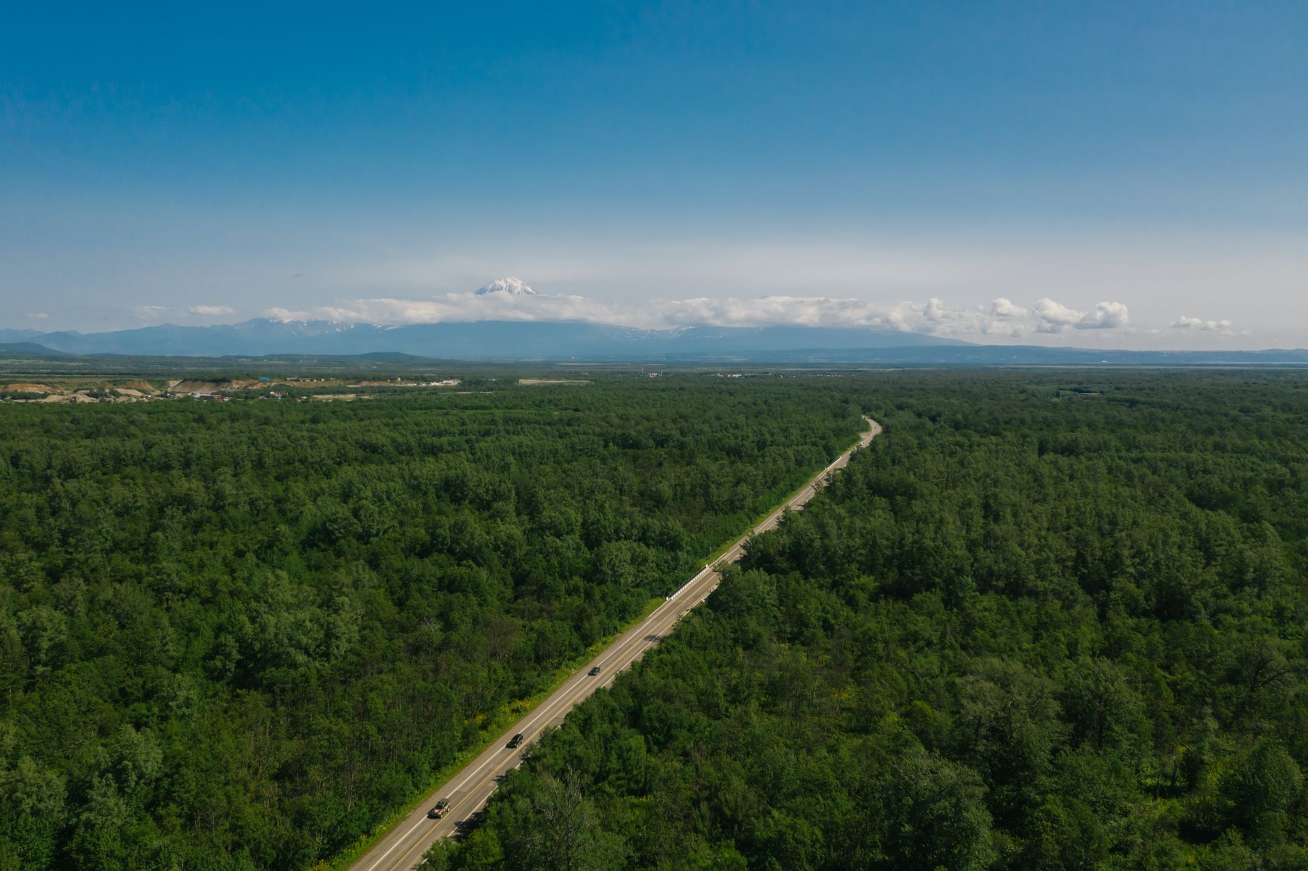 Drone shot capturing a road cutting through lush green forest with distant mountains.