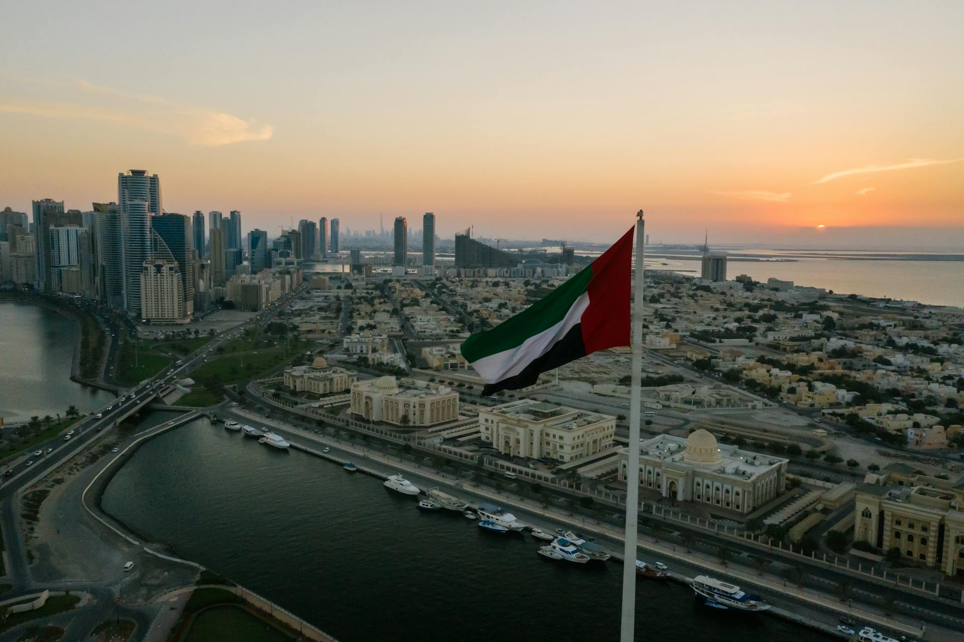 A breathtaking aerial view of a UAE city skyline with a prominent national flag at sunset, showcasing urban architecture.