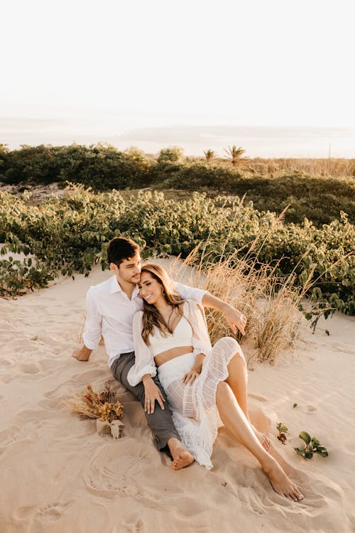 Full body of young man and woman cuddling on sand near bushes during date